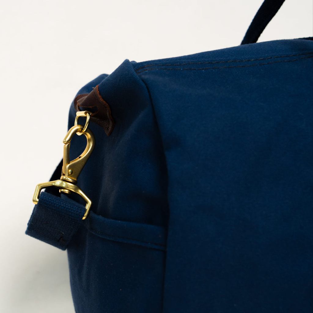 Close-up of a brass shoulder-strap clasp and leather trim on a midnight-navy Steele Canvas bag.