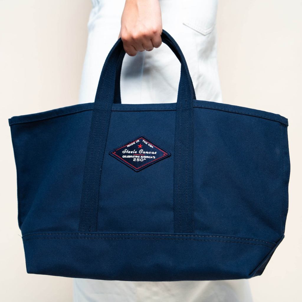 Person holding a midnight-navy Steele Canvas tote with short handles and the 250th anniversary patch.