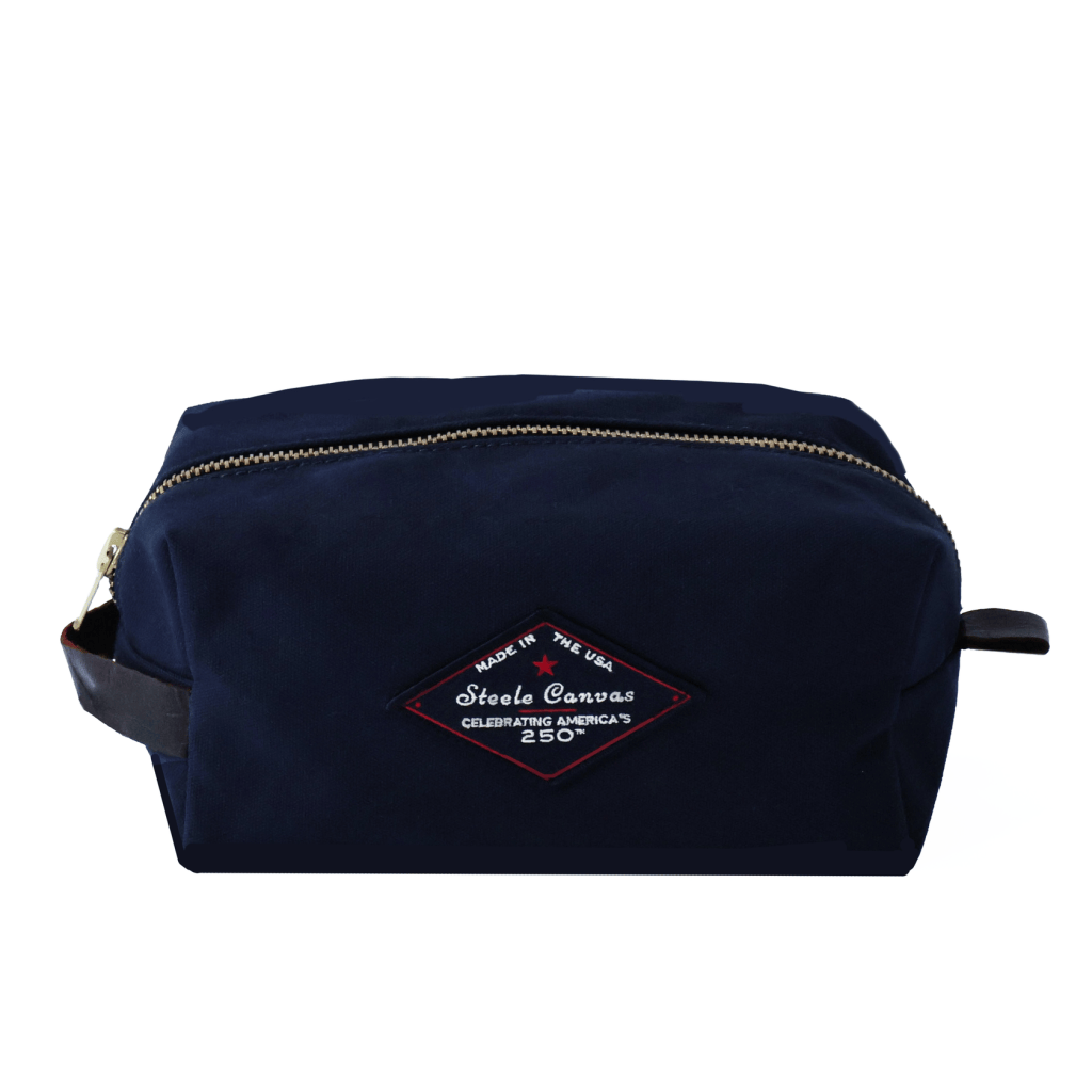Front view of a midnight-navy Steele Canvas Dopp kit with brass zipper, leather side handle, and 250th anniversary patch.