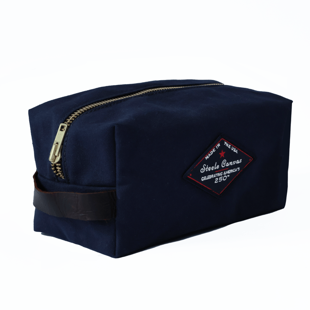 Angled view of a midnight-navy Steele Canvas Dopp kit with brass zipper, leather handle, and 250th anniversary patch.